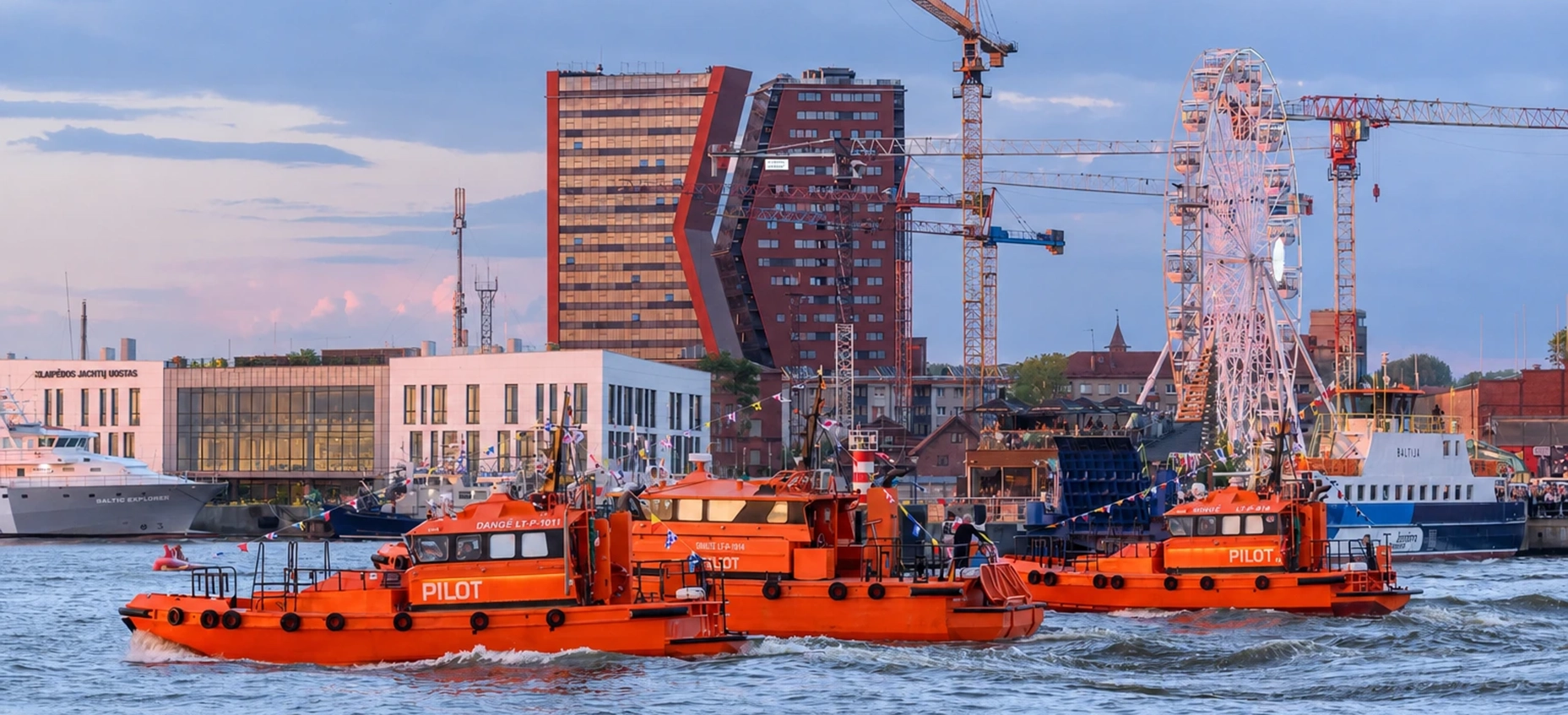 Orange pilot boats in Klaipėda harbor with cranes, ferris wheel, and waterfront buildings.